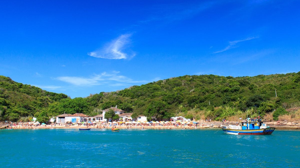 Praia movimentada com mar azul-turquesa, pessoas, guarda-sóis, casas brancas, barco e colinas verdes.