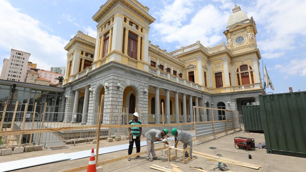 Três operários trabalham em canteiro de obras, com edifício histórico amarelo e cinza ao fundo
