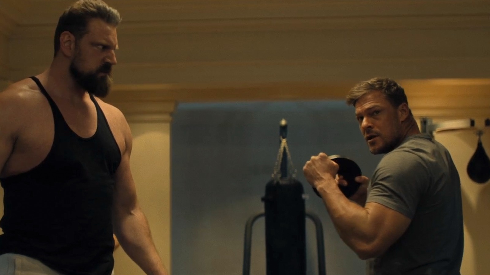 Alan Ritchson, o protagonista de Reacher, em cena da série que tem a quarta temporada confirmada para agosto de 2026. Dois homens musculosos em uma academia, um com barba em camiseta preta e outro com peso.