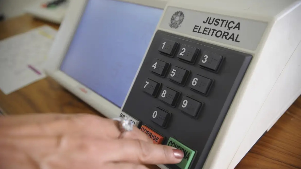 Eleitores devem ficar atentos às mudanças nos locais de votação e confirmar sua seção eleitoral antes do dia do pleito. Mão pressionando o botão 'CONFIRMA' de uma urna eletrônica com 'JUSTIÇA ELEITORAL' visível
