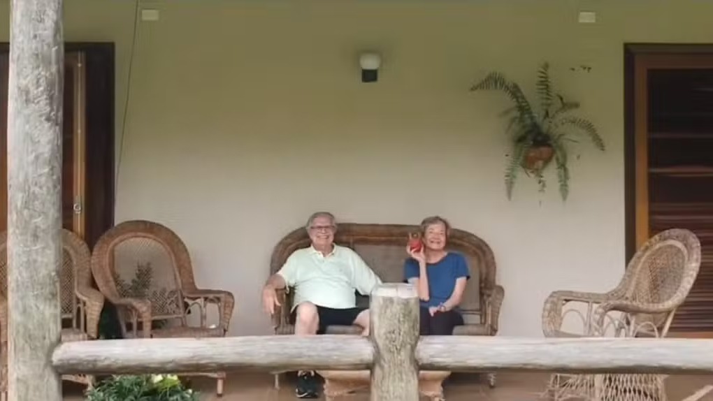 Homem e mulher idosos sorrindo em banco de vime na varanda, mulher segura celular