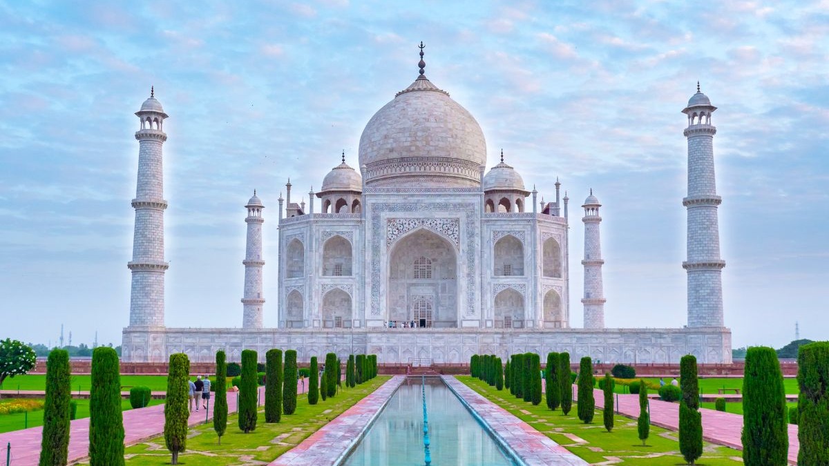 O Taj Mahal, suntuoso mausoléu de mármore branco na Índia, figura entre as Sete Maravilhas do Mundo Moderno. Taj Mahal branco com cúpulas, minaretes, lago refletor e jardins bem cuidados sob céu azul.