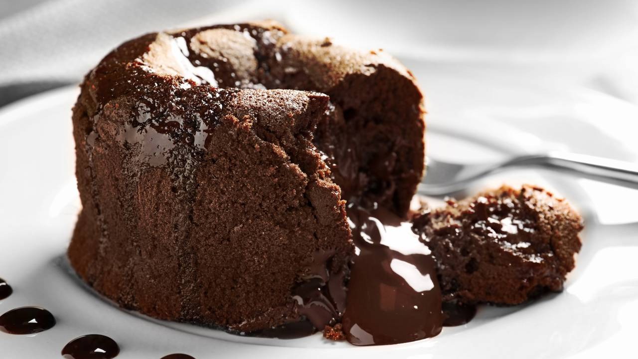 O delicioso petit gâteau, um pequeno bolo com o irresistível recheio cremoso de chocolate, conforme a receita. Petit gâteau de chocolate meio aberto com recheio derretido escorrendo sobre prato branco