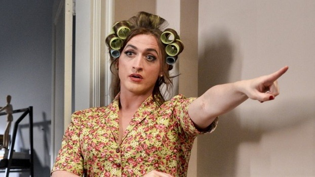 Pessoa caracterizada como Dona Herm&iacute;nia, com bobes no cabelo e camisa floral, apontando &agrave; direita.