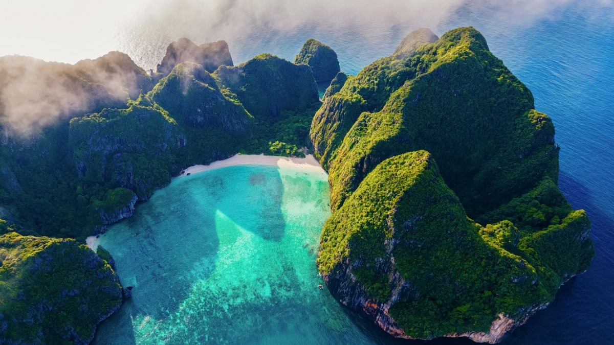 Vista a&eacute;rea de uma ba&iacute;a tropical com &aacute;gua turquesa, praia de areia branca e montanhas verdes.