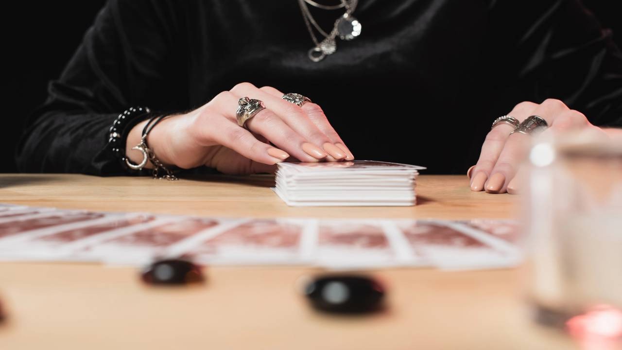 Mãos sobre cartas oraculares, remetendo à busca por compreender tendências e a influência cósmica, tema central da astrologia para a sorte. Mãos femininas com anéis e pulseiras tocam um baralho de cartas sobre uma mesa de madeira.