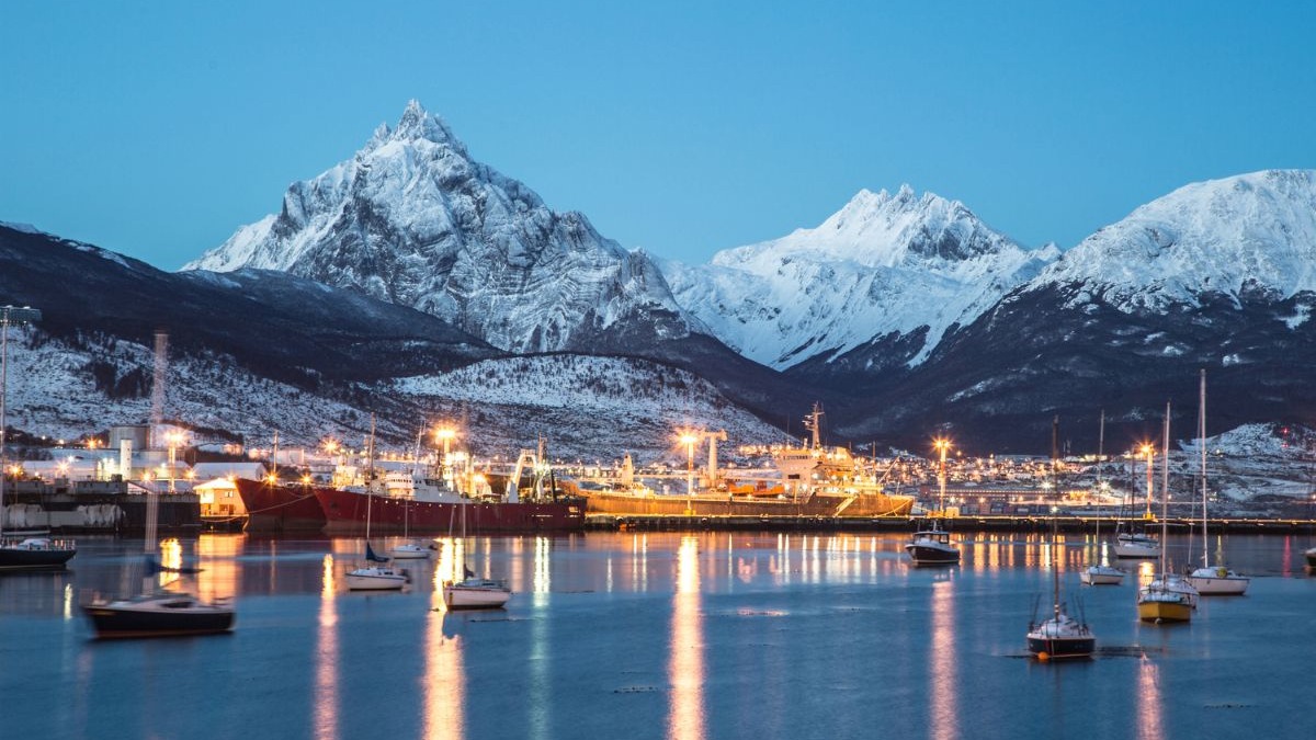 Ushuaia, na Argentina, um dos destinos do Sul do continente que se torna mais acessível com a valorização do real. Cidade costeira com porto e montanhas nevadas ao entardecer, luzes urbanas refletem na água, barcos.