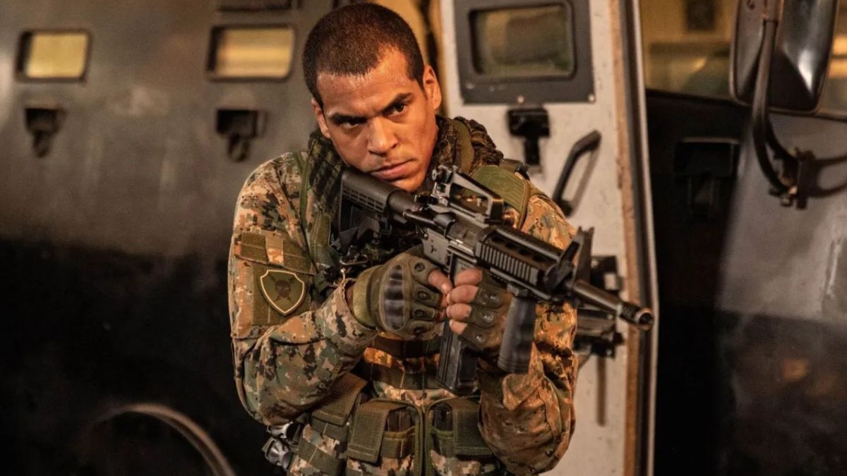 Homem em uniforme camuflado, segurando fuzil, luvas táticas, olhar sério. Fundo escuro.