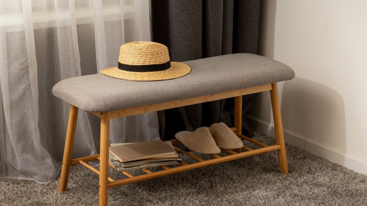 Banco cinza acolchoado com chap&eacute;u de palha, chinelos e livros em prateleira inferior, sobre tapete.