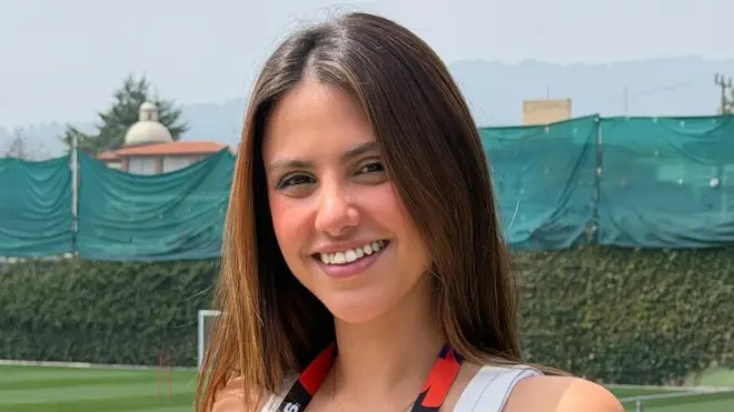 Victória Leite, que cobrirá o futebol inglês, é a nova correspondente da TNT Sports na Inglaterra. Foto: Reprodução Mulher sorrindo, cabelo castanho, blusa branca, com campo de futebol e grade verde ao fundo