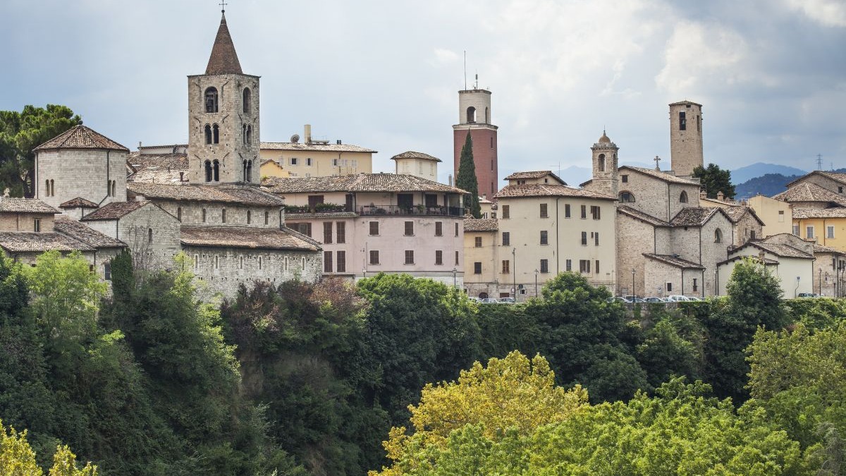 A elegância do centro histórico de Ascoli Piceno, em mármore travertino, revela o charme das cidades italianas acessíveis. Cidade histórica italiana com edifícios de pedra e torres sineiras, em colina verde.