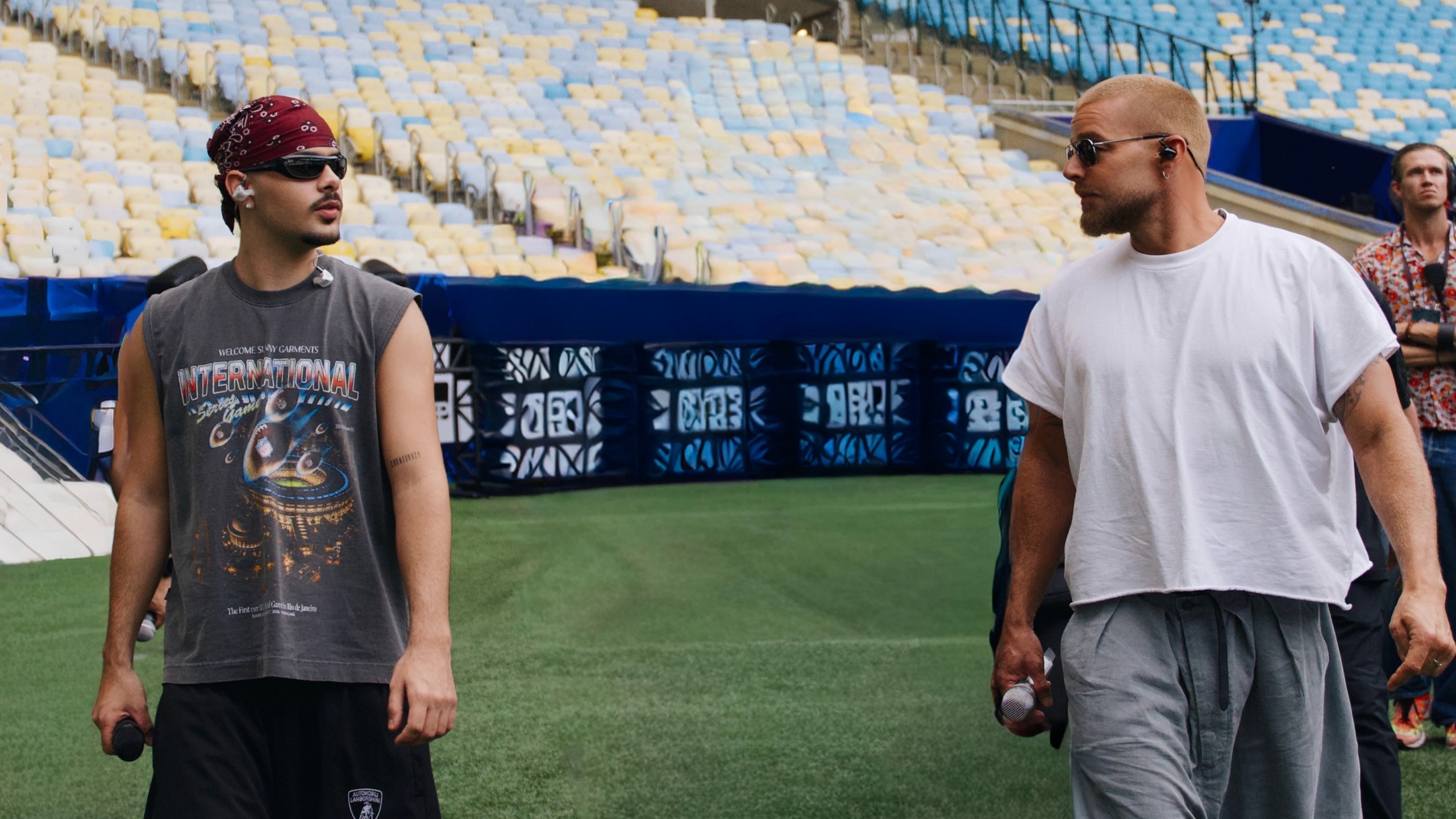 Pedro Sampaio (esq.) e Ricky Martin se preparam para o show do intervalo do jogo da NFL no Maracanã. Dois homens com microfones em campo de estádio. Um veste bandana e camiseta "INTERNATIONAL"