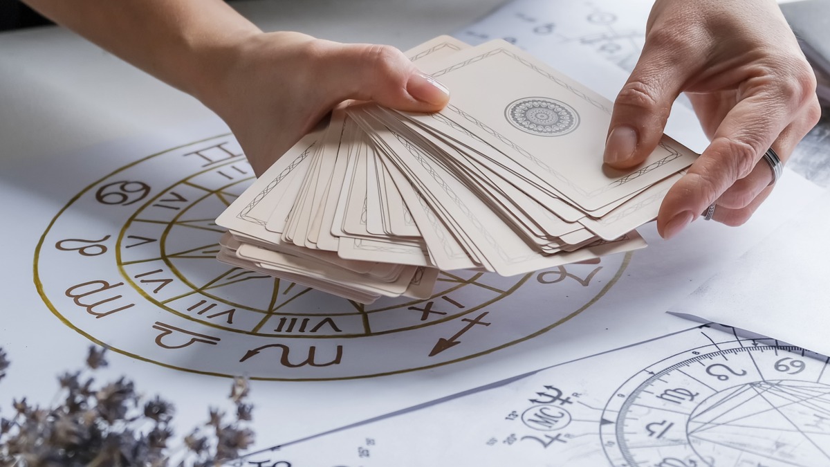 Ferramentas esotéricas como cartas e mapas astrais ilustram a busca por respostas sobre o futuro dos relacionamentos em 2026, segundo a astrologia. Mãos seguram baralho de cartas sobre mapas astrais com signos e símbolos astrológicos.
