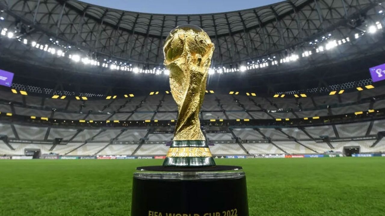 O cobiçado troféu da Copa do Mundo, o grande objetivo que Marquinhos busca com a Seleção Brasileira para entrar na história. Troféu dourado da Copa do Mundo 2022 em pedestal preto, no centro de um estádio de futebol vazio.