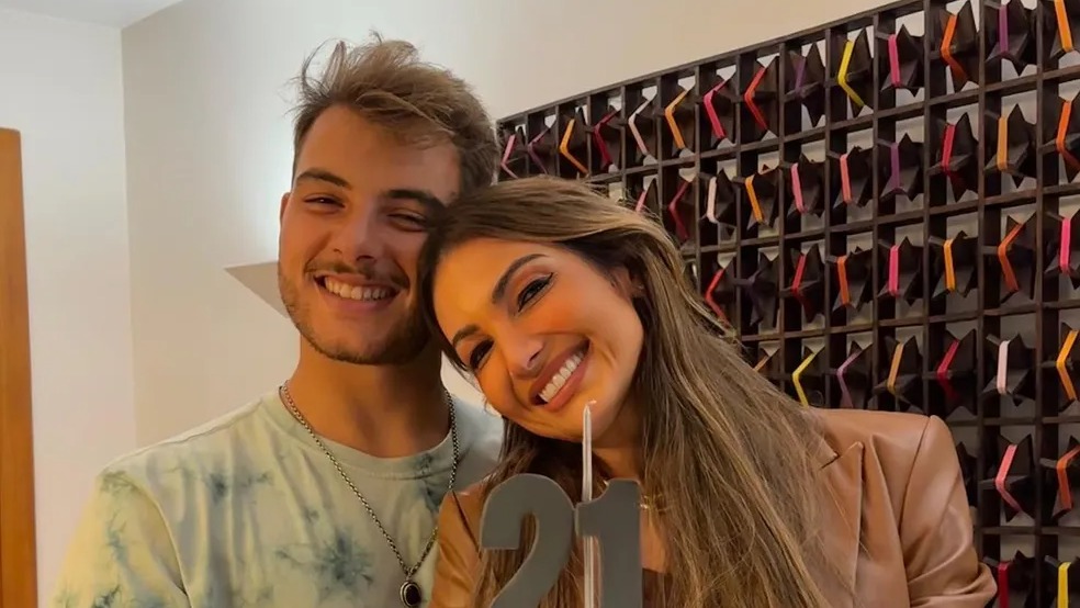 Jovens talentos, herdeiros de grandes nomes, celebram novas fases em suas vidas e carreiras. Um homem e uma mulher sorrindo, ela segura o número 21, parede decorada ao fundo