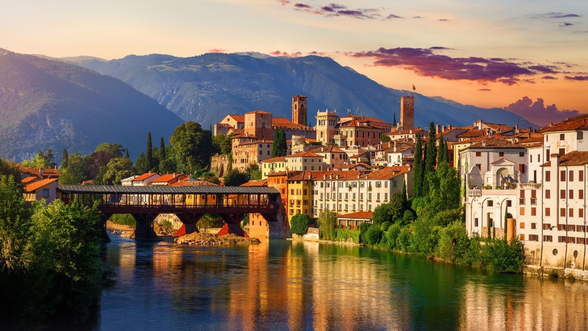 Bassano del Grappa, com sua icônica Ponte degli Alpini, destaca-se como opção charmosa para quem busca nova vida na Itália. Cidade italiana com ponte de madeira coberta sobre rio e montanhas, ao pôr do sol.
