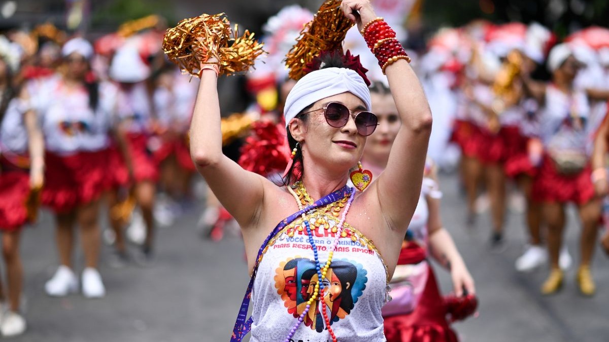 Mulher com turbante branco, &oacute;culos de sol, pompons dourados e blusa estampada em bloco de Carnaval.