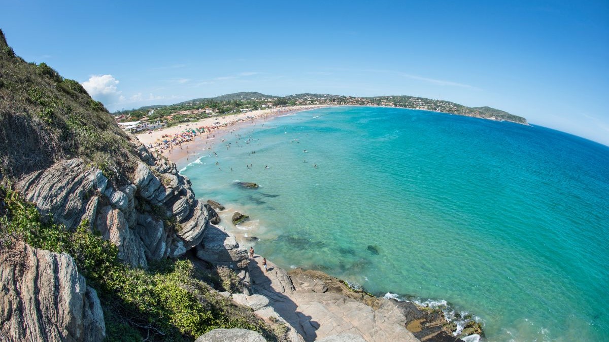 Vista panorâmica de praia movimentada com mar azul-turquesa, rochas e vegetação sob céu claro.