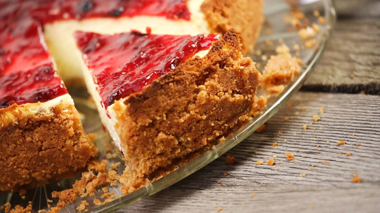 Aprenda a fazer um cheesecake de frutas vermelhas com base crocante, recheio cremoso e cobertura caseira. Fatia de cheesecake de frutas vermelhas com base crocante, creme branco, em prato de vidro