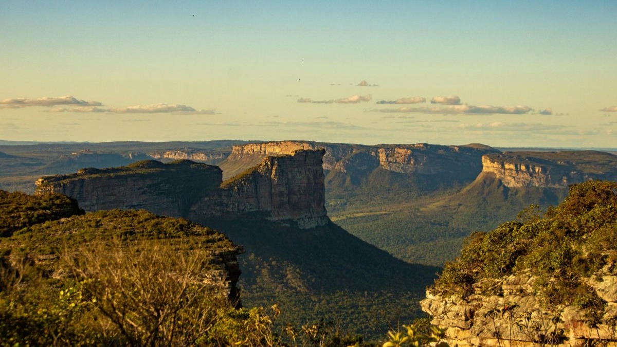 Vasta paisagem montanhosa com platôs rochosos e vales verdes sob um céu azul.