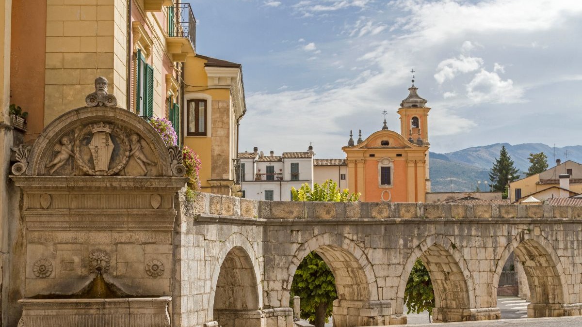 Sulmona, no coração da Itália, oferece uma rica história e arquitetura em seu centro medieval bem preservado. Fonte de pedra ornamentada com arcos, edifícios históricos e uma igreja em Sulmona, Itália.