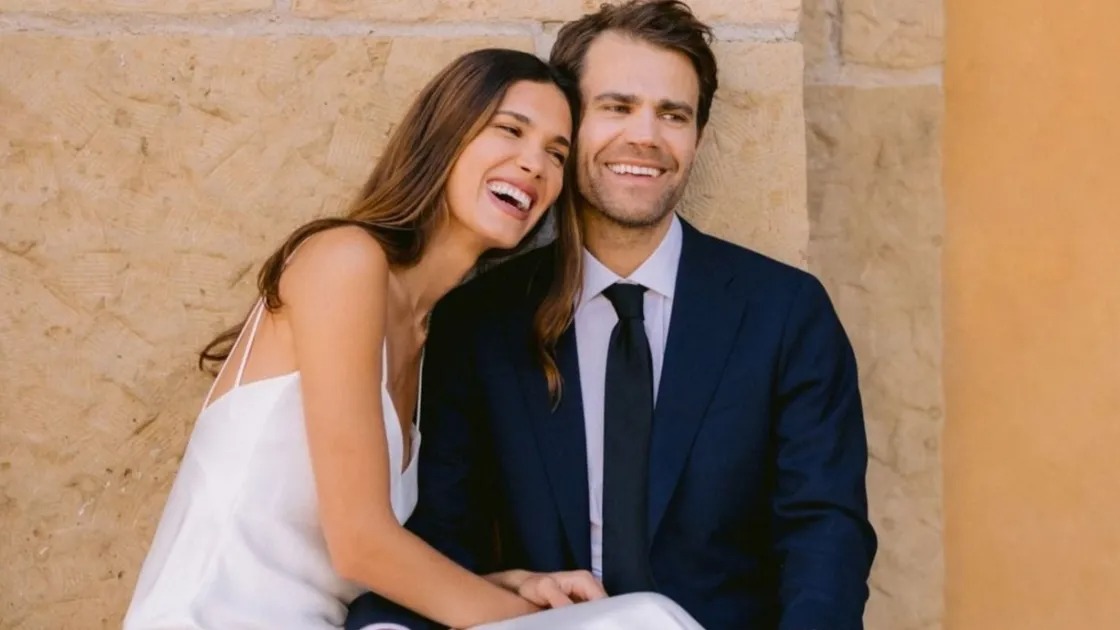 Casal sorridente, Paul Wesley e Natalie Kuckenburg, abra&ccedil;ados, ela em vestido branco, ele de terno