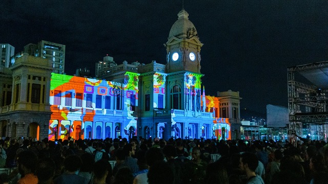 Prédio histórico com projeções coloridas, torre do relógio, multidão em evento noturno