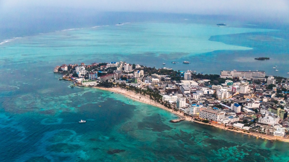 Vista a&eacute;rea de cidade costeira com edif&iacute;cios, praia de areia, palmeiras e mar azul-turquesa com barcos.