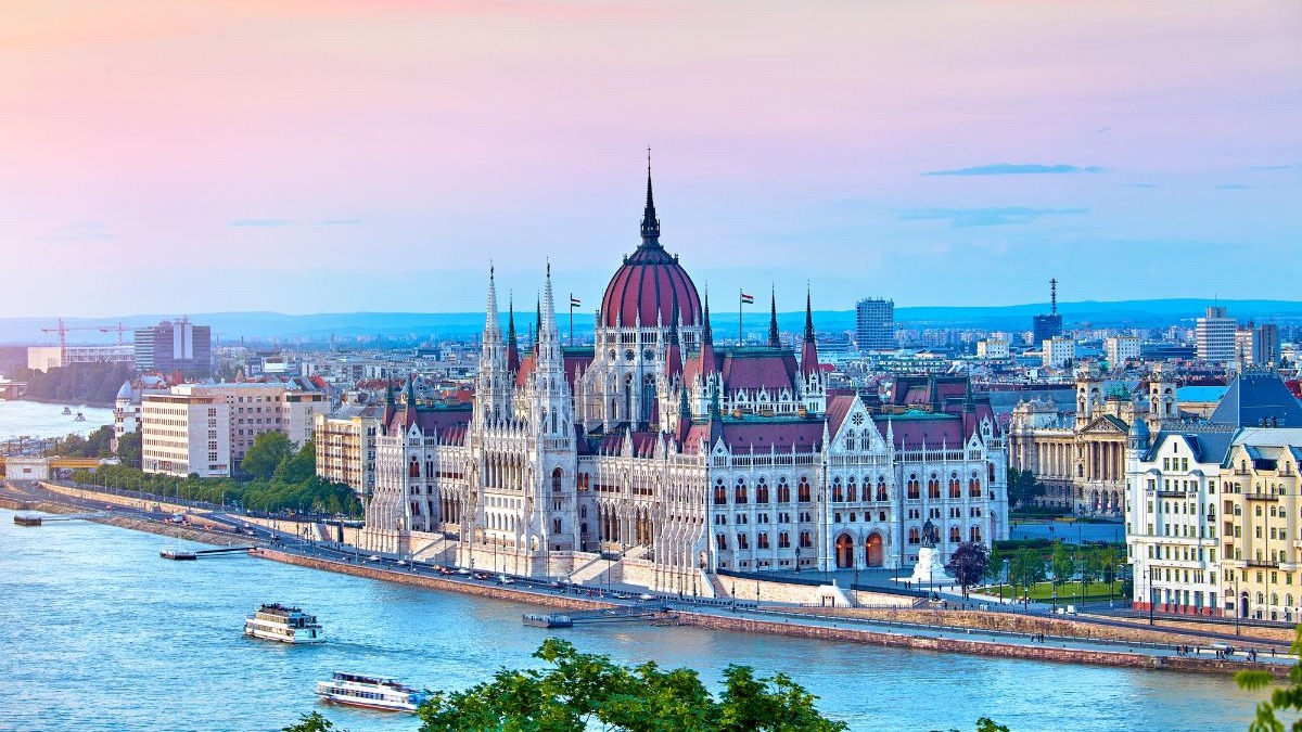 Imponente Parlamento de Budapeste junto ao rio Dan&uacute;bio, com barcos e c&eacute;u rosado.