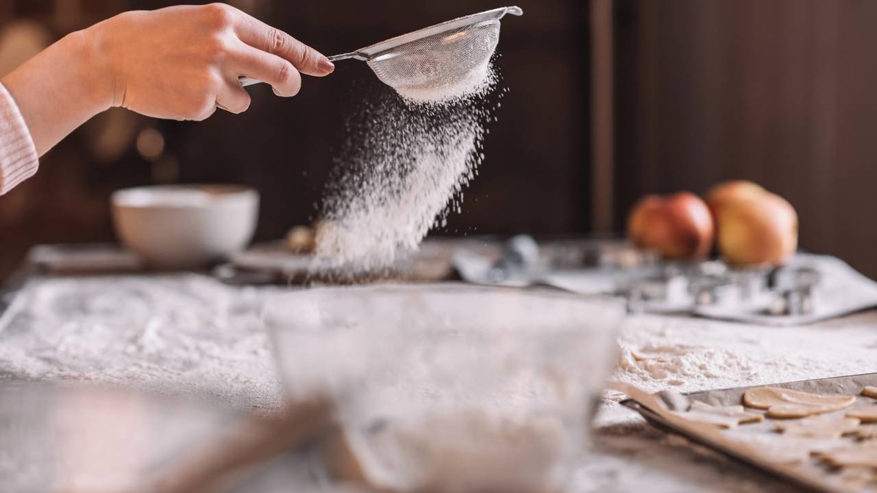 A preparação cuidadosa dos doces é a base para o sucesso das vendas, facilitadas por métodos de pagamento como o Pix. Mão peneira farinha branca sobre mesa, com tigela e formas de massa no fundo.