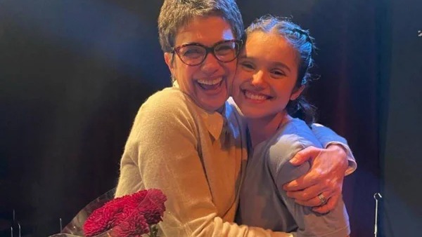 Sandra Annenberg celebra a carreira teatral da filha Elisa Annenberg-Paglia, que brilha nos palcos após formação em Nova York. Duas mulheres sorrindo se abraçam. Uma de óculos, cabelo grisalho. Outra com tranças. Flores vermelhas.