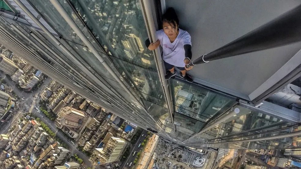 A busca por visualizações levava influenciadores como Wu Yongning a escalar arranha-céus, uma prática perigosa que resultou em tragédia. Pessoa em beiral de arranha-céu com selfie stick e vista aérea da cidade.