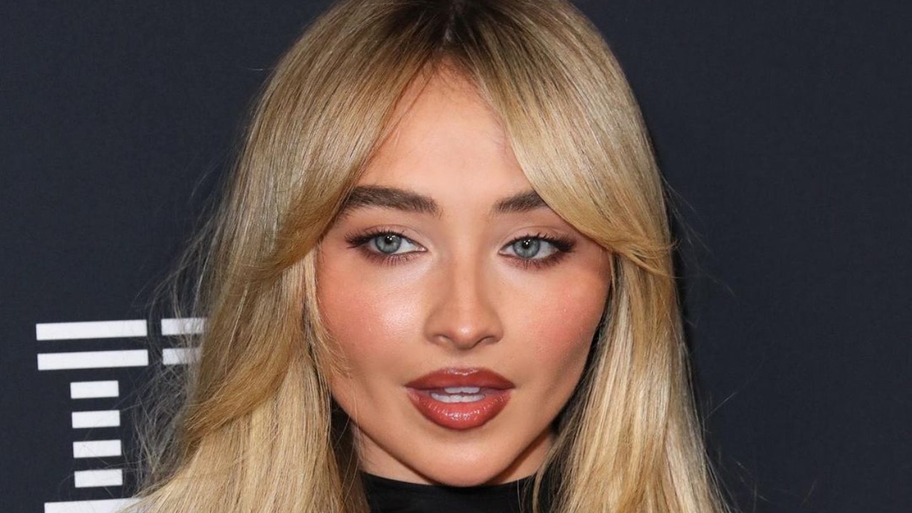 Com camadas que emolduram o rosto, o Butterfly Cut é um dos cortes em alta para quem busca volume e movimento sem sacrificar o comprimento. Close-up de mulher loira com cabelo em camadas, olhos azuis, batom marrom avermelhado.