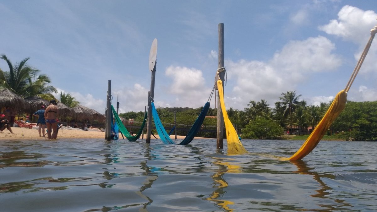 Redes coloridas na &aacute;gua, praia tropical com coqueiros, pessoas e c&eacute;u azul com nuvens.