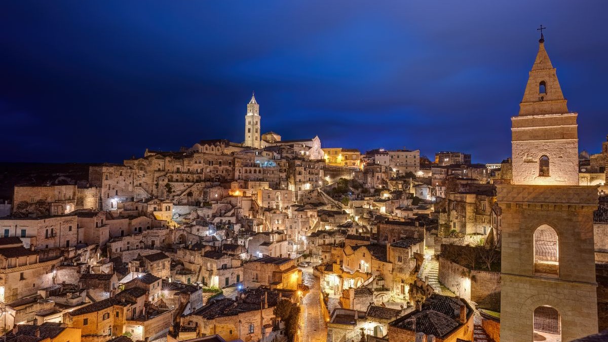 Matera, a cidade de pedra, revela seu visual dramático e milenar à noite, um espetáculo à parte na Itália. Matera, Itália, à noite, com edifícios de pedra iluminados e torres de igreja.