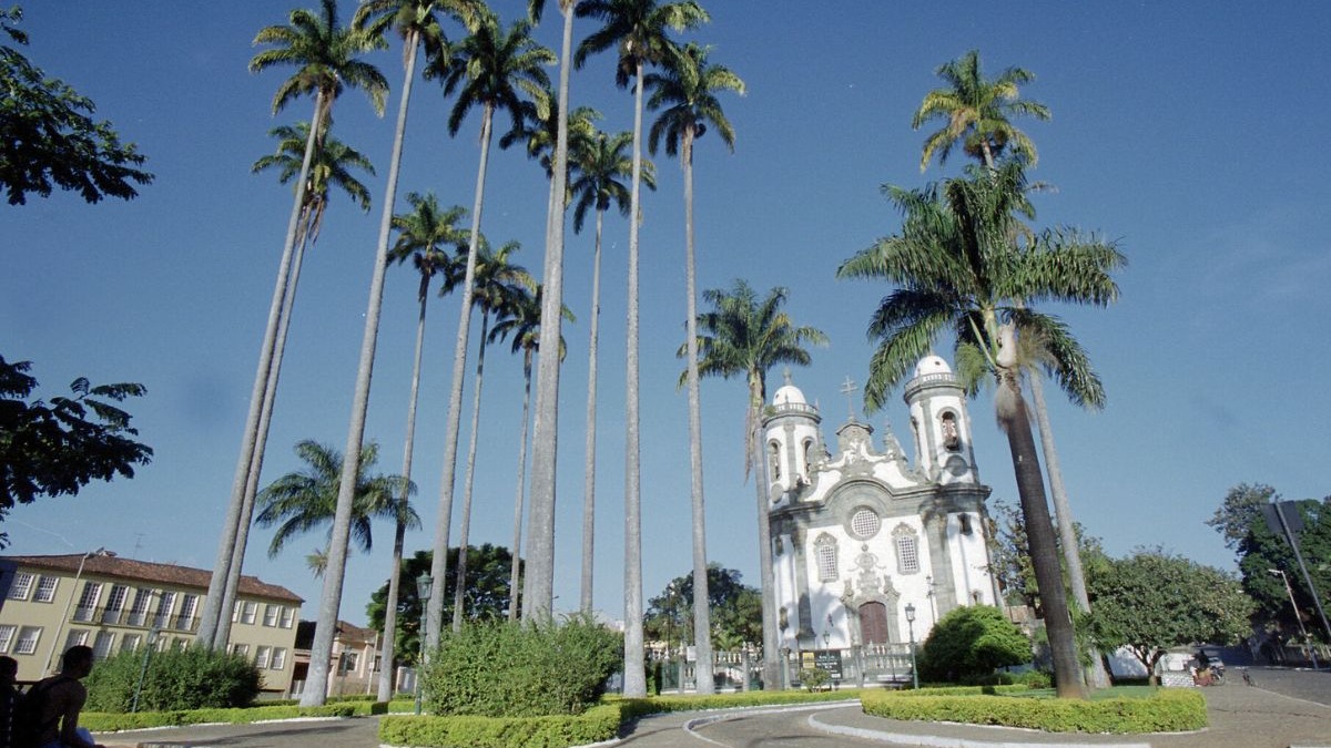 Igreja barroca branca com duas torres, rodeada por altas palmeiras imperiais sob c&eacute;u azul.