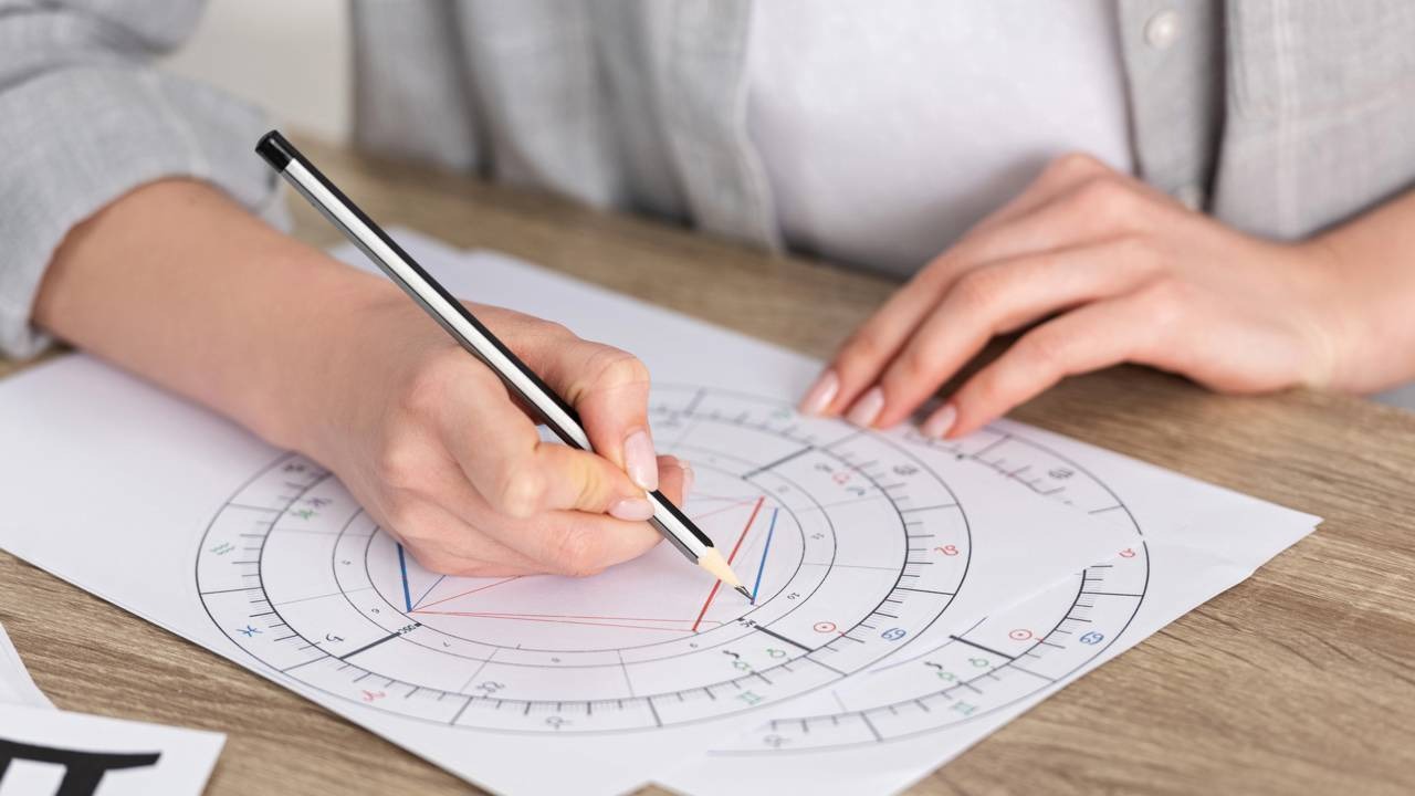 Entenda como os trânsitos planetários são interpretados para revelar o futuro amoroso dos librianos no segundo semestre de 2026. Mãos desenhando com lápis em um mapa astral circular sobre papel branco em mesa de madeira.