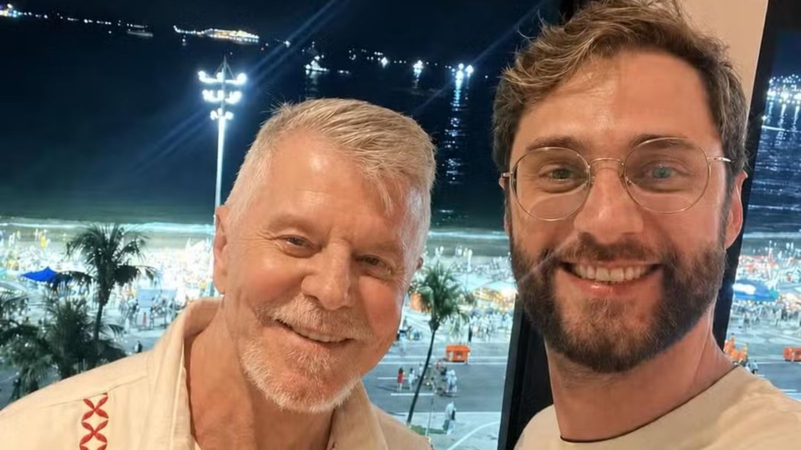 Miguel Falabella e um jovem sorrindo em selfie noturna com praia iluminada ao fundo
