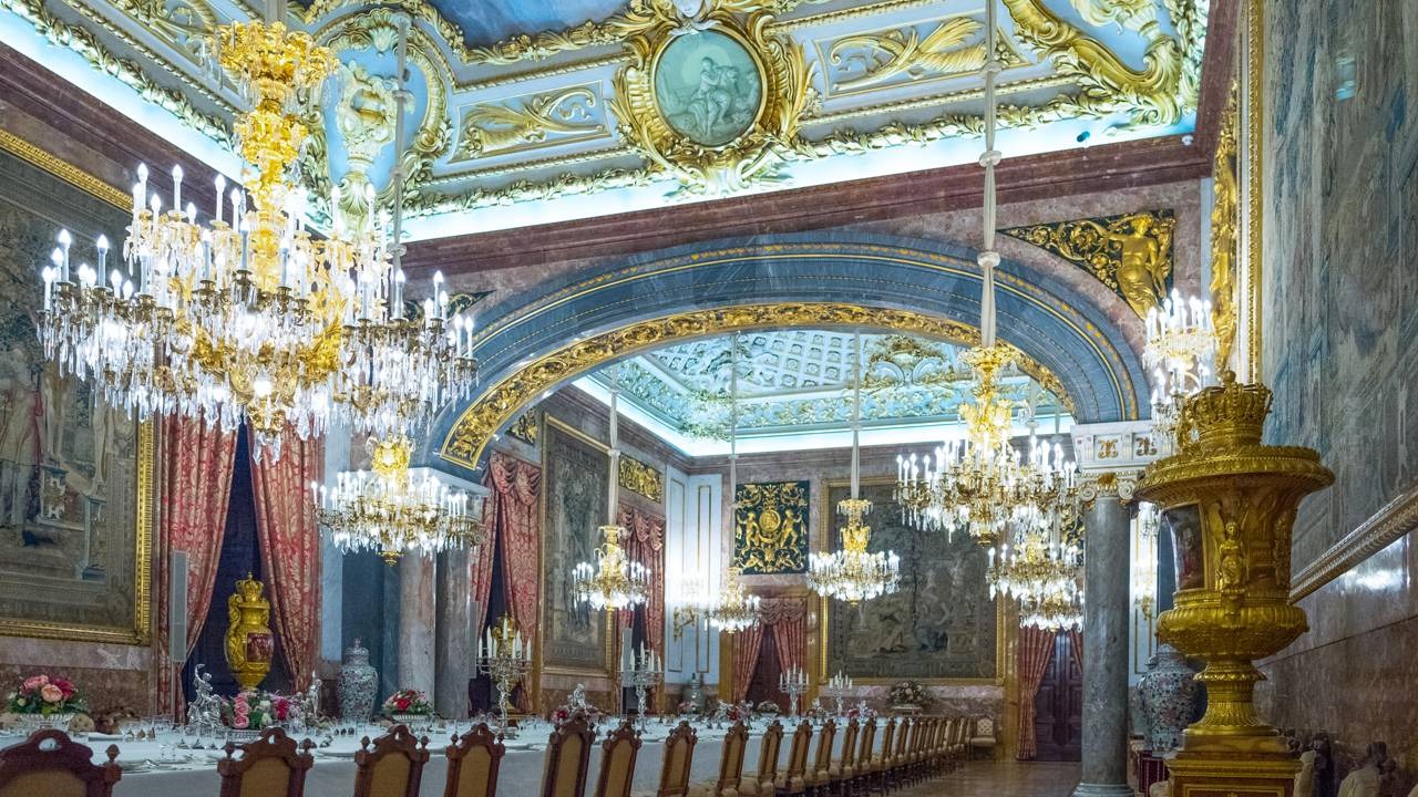 A Sala de Jantar de Gala do Palácio Real de Madrid, com seus lustres monumentais, é um dos espaços que recebe cuidados especiais de restauração Opulento salão de jantar real com lustres de cristal, teto dourado e azul, e longa mesa posta