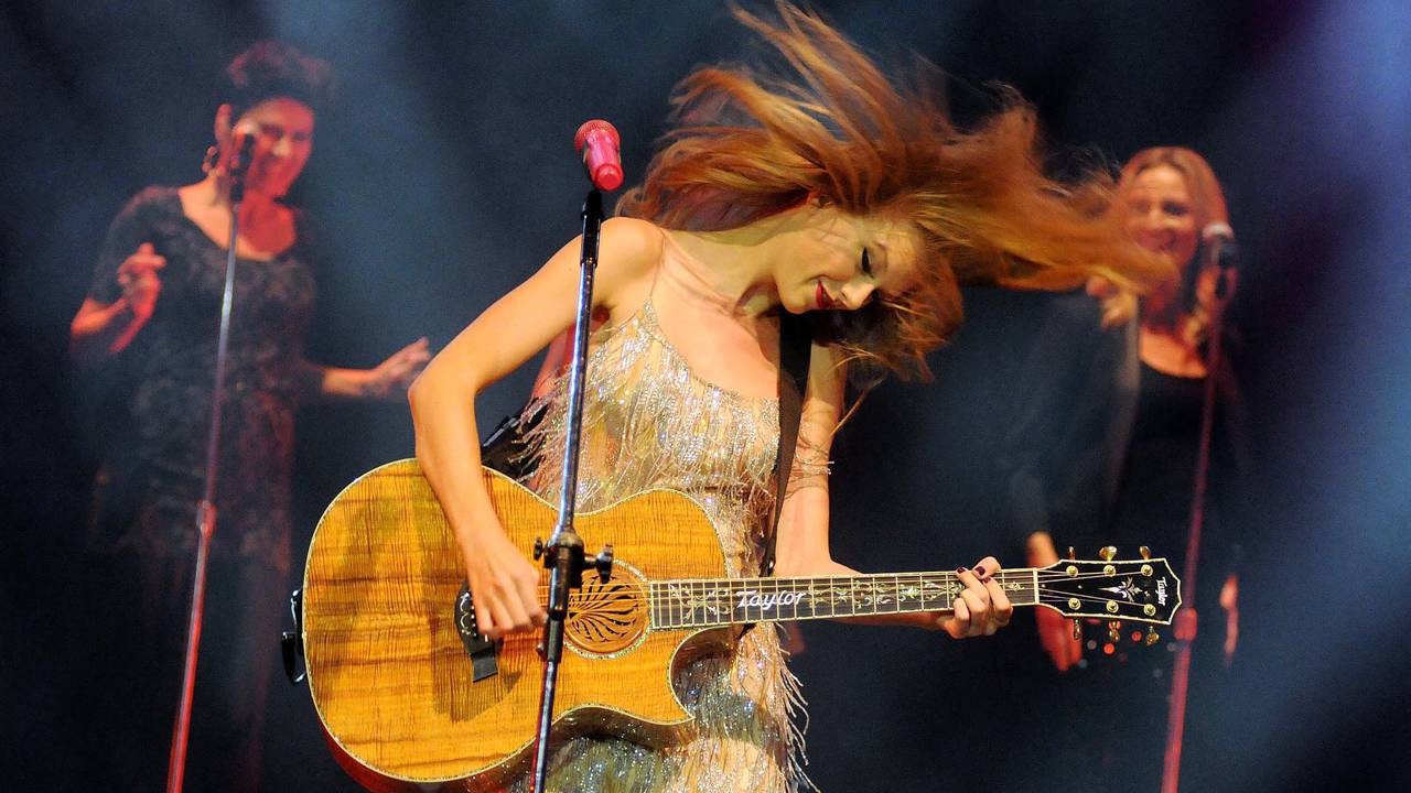 A performance energética de Taylor Swift em palco, ilustrando a ambição sagitariana de levar sua música a milhões de fãs. Taylor Swift tocando violão personalizado 'Taylor' em palco, cabelo ruivo voando.