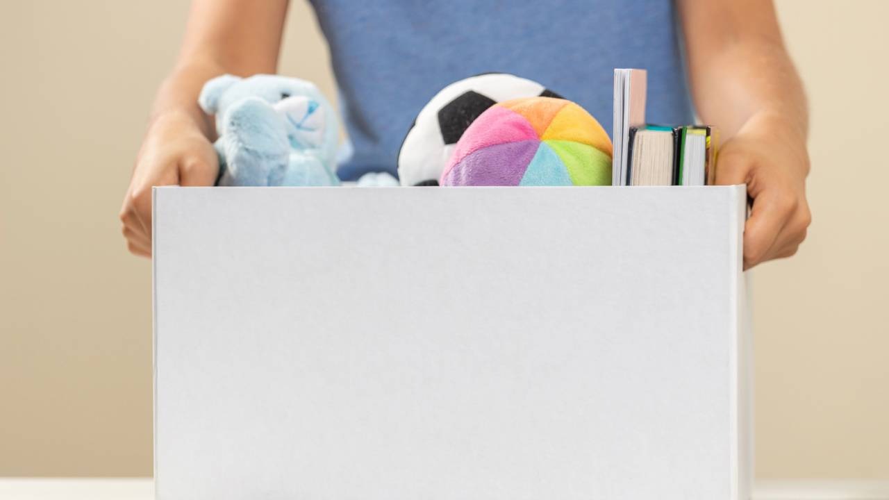 Organizar por categoria, como os itens guardados nesta caixa, é essencial para o método KonMari de Marie Kondo Pessoa segurando uma caixa branca cheia de brinquedos de pelúcia coloridos e livros pequenos