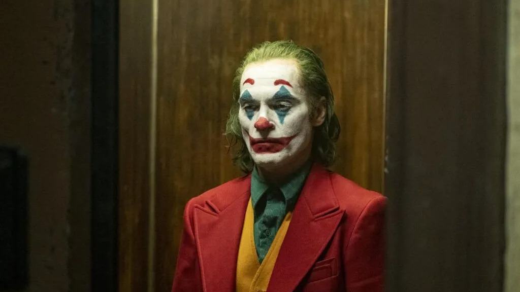 Joaquin Phoenix como Coringa, com maquiagem branca, vermelha e azul no rosto e cabelo esverdeado. Ele usa um paletó vermelho, camisa verde e colete amarelo, com expressão introspectiva. O ator está em um ambiente interno com paredes de madeira escuras.