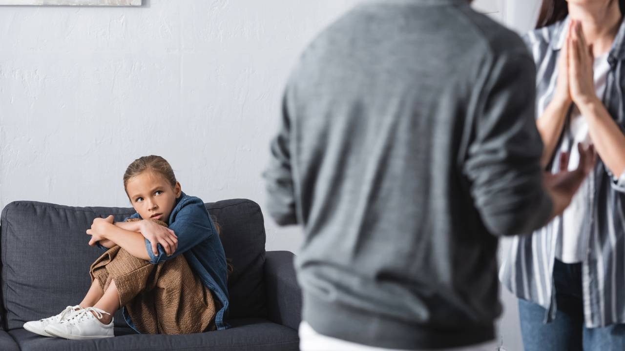 A hipervigilância surge como mecanismo de defesa em crianças expostas a ambientes familiares imprevisíveis e conflituosos Menina, braços cruzados no sofá, olha adultos desfocados interagindo com tensão