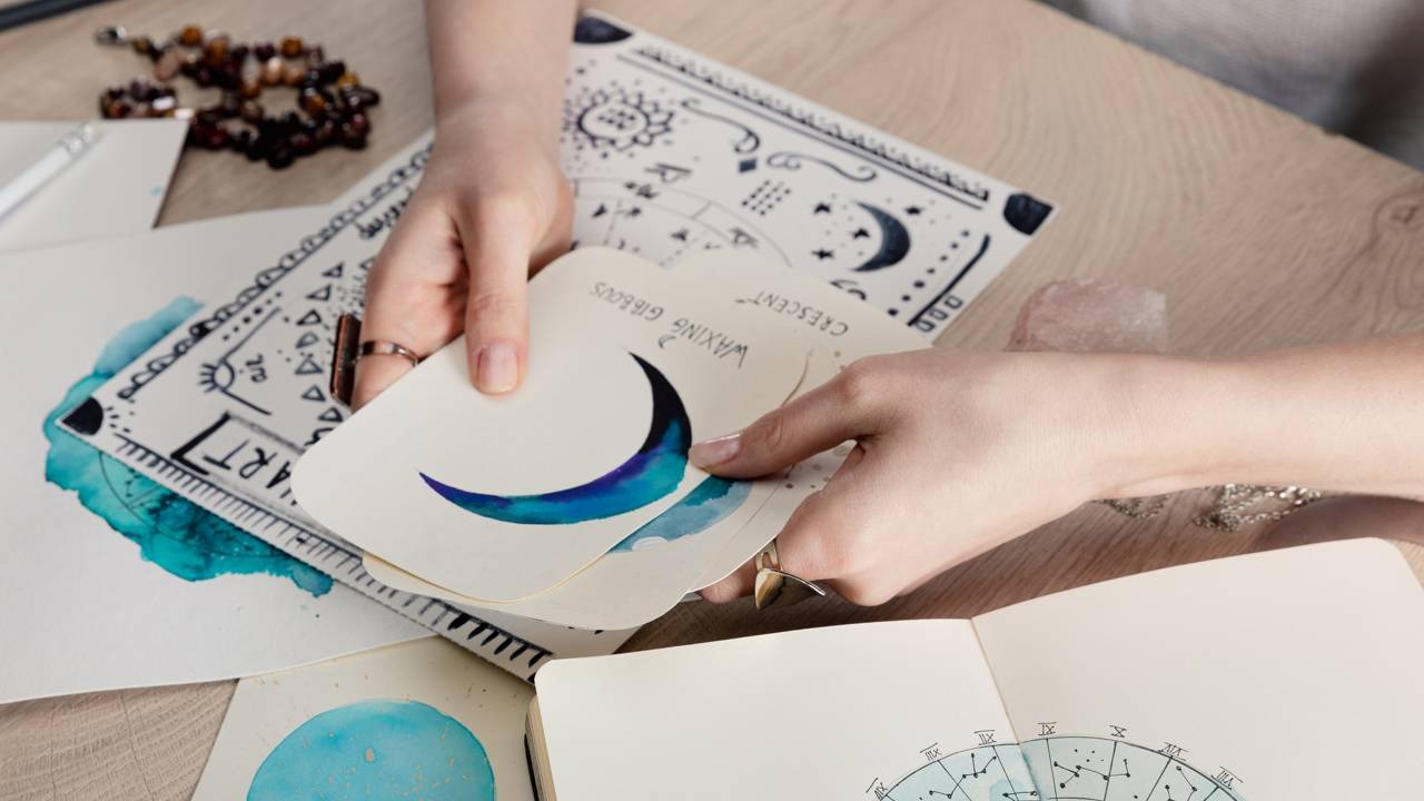 Ilustrações astrológicas e criatividade artística se alinham, sugerindo a conexão entre os signos e o sucesso digital Mãos seguram cartas com fases da lua em aquarela, sobre mesa com desenhos astrológicos