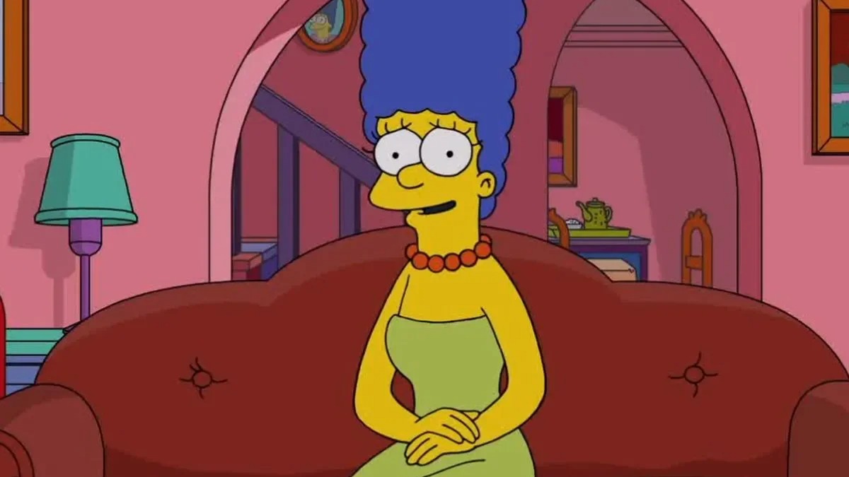 Marge Simpson, de cabelo azul, vestido verde, sentada no sof&aacute; vermelho com express&atilde;o pensativa.