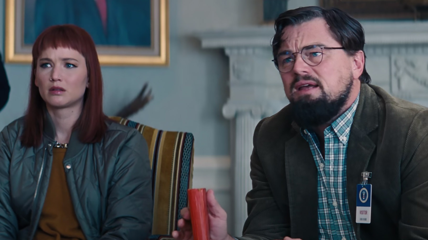 Leonardo DiCaprio e Jennifer Lawrence no filme "Não Olhe Para Cima". Jennifer Lawrence e Leonardo DiCaprio, ele de óculos e barba, com crachá e objeto vermelho na mão