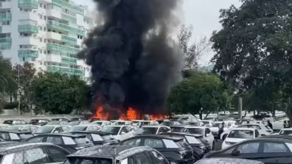 Incêndio atinge veículos elétricos após colisão de helicópteros no Recreio dos Bandeirantes Grande incêndio com fumaça preta densa em estacionamento com vários carros e edifício