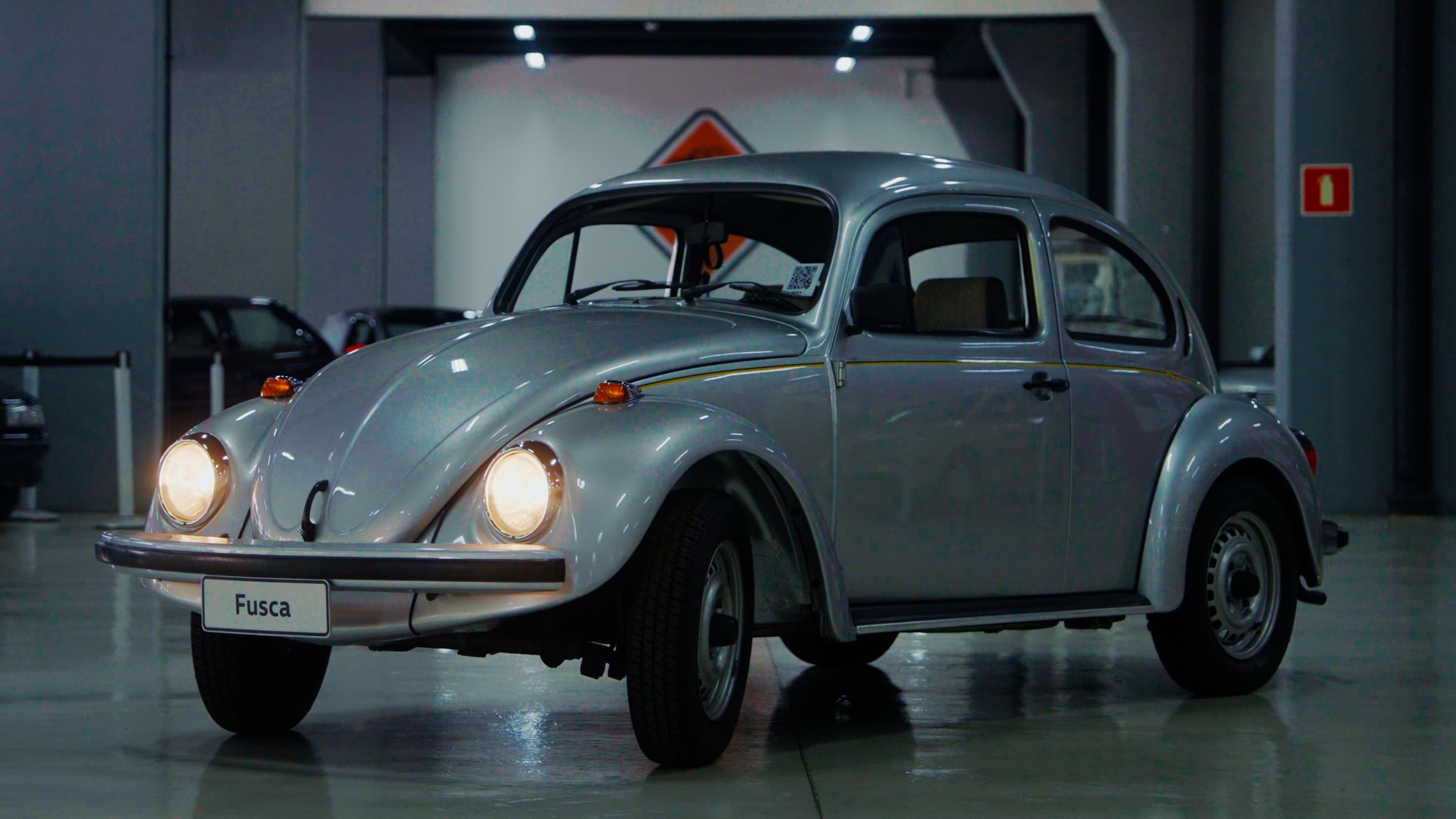 Cl&aacute;ssico Volkswagen Fusca prateado com far&oacute;is acesos e placa 'Fusca' em showroom.