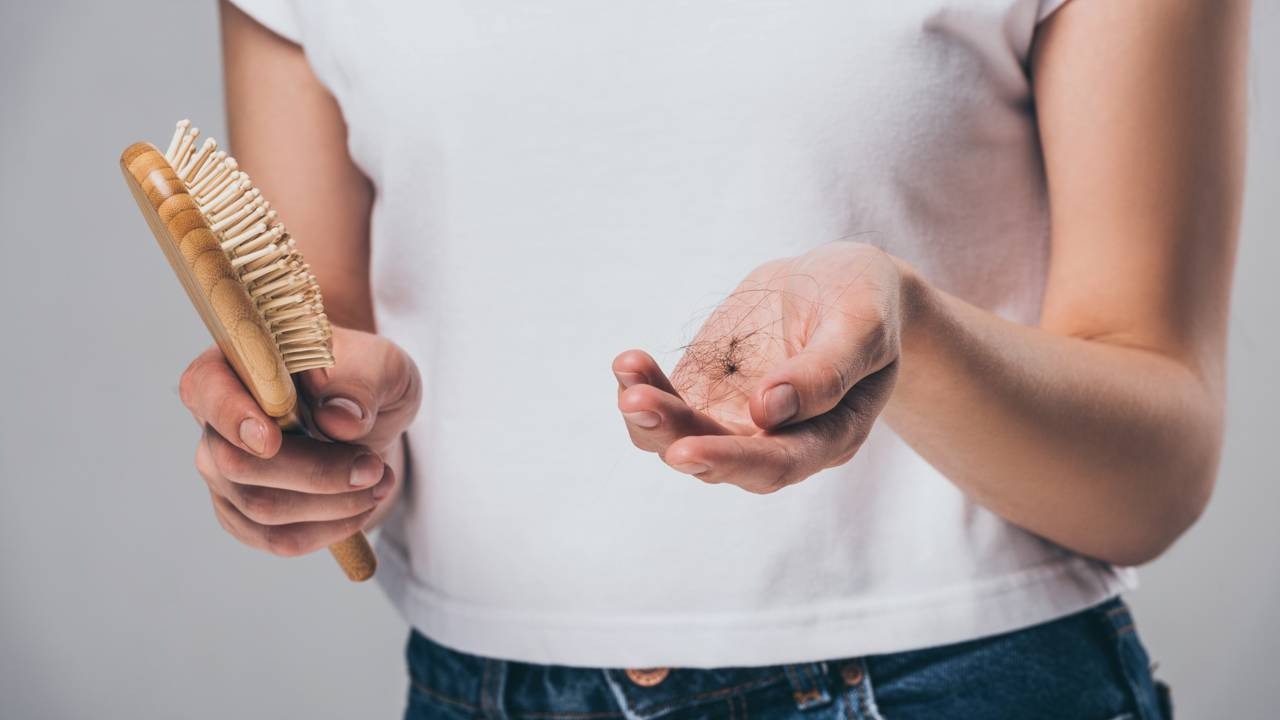 A queda de cabelo, sintoma comum da alopecia, é uma condição de saúde que afeta milhões, discutida em detalhes nesta matéria. Mãos seguram escova de madeira em uma mão e punhado de cabelos soltos na outra.