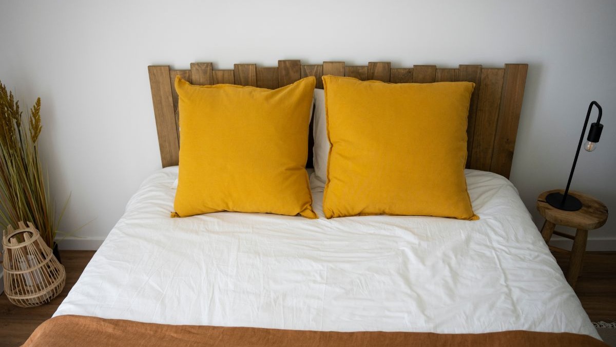 Cabeceiras de madeira, como a da imagem, harmonizam com a tendência de 2026 que valoriza o toque natural e a sustentabilidade nos quartos. Cama com cabeceira de madeira, almofadas amarelas, roupa de cama branca e detalhes naturais.